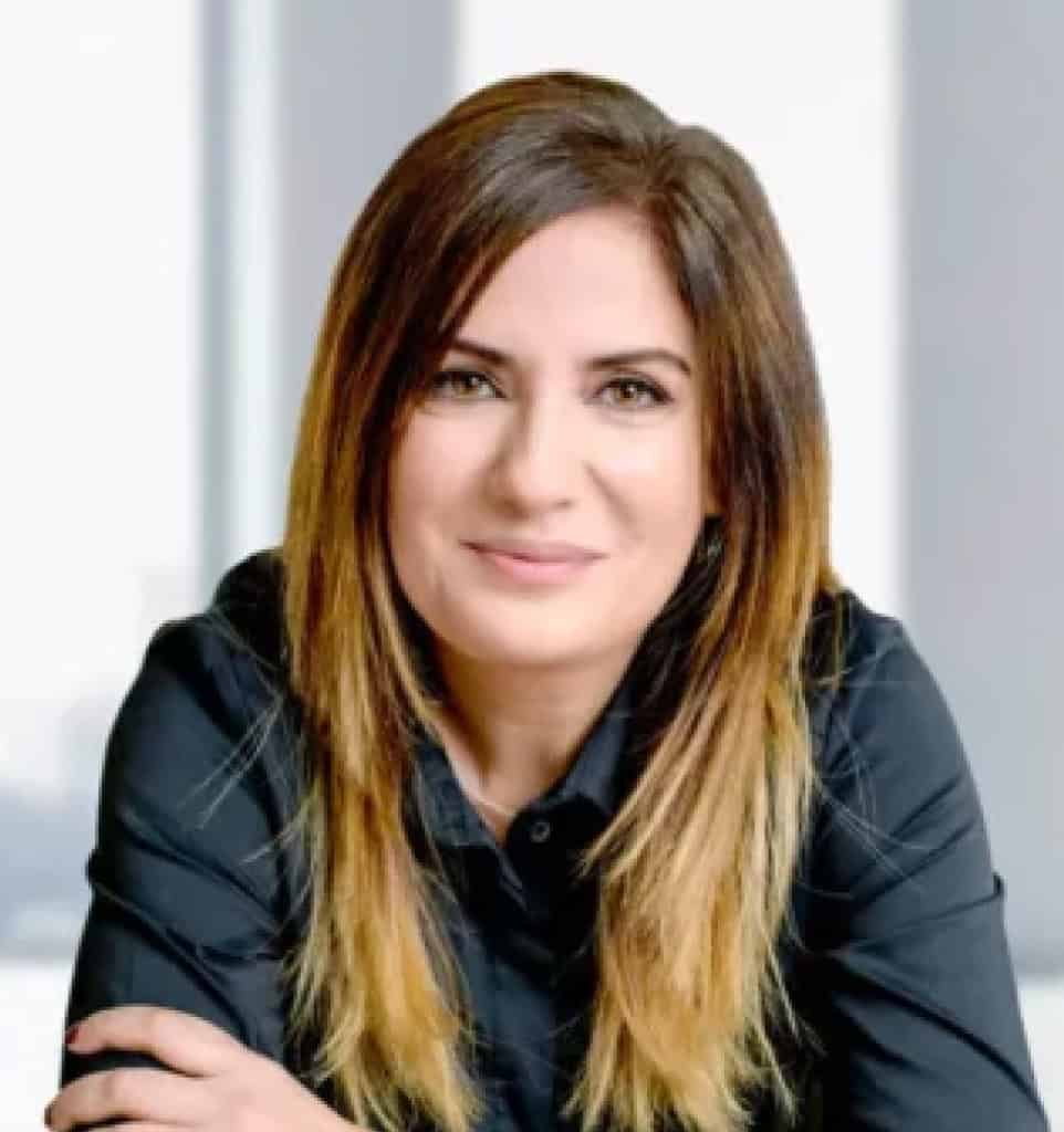 אישה עם שיער חום ישר באורך הכתפיים וקצוות בלונדיניים, לבושה חולצה שחורה, יושבת מול רקע מטושטש עם חלונות גדולים. דגנית אלישיב, עורכת דין ומגשרת.