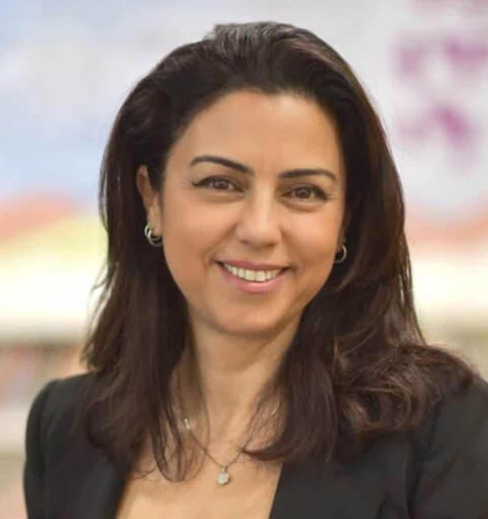 אישה עם שיער כהה, לבושה בז'קט שחור ועונדת שרשרת, מחייכת למצלמה בתוך חלל סגור, ומשדרת ביטחון עצמי כשהיא מדברת על איזון נטו במשאבים פיננסיים.