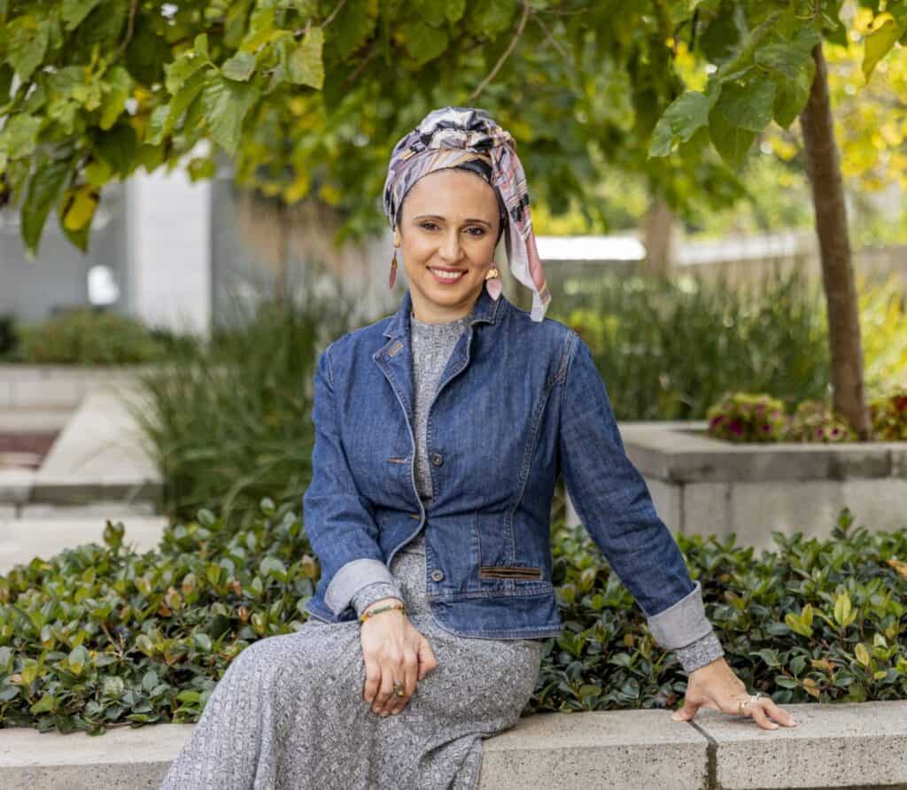 אישה חבושה במטפחת בדוגמה, לבושה ז'קט ג'ינס ושמלה ארוכה, יושבת על ספסל בטון בגינה חיצונית, מחייכת למצלמה ומקרינה את החום של ליווי רגשי מאת אילנה איילה גבאי.