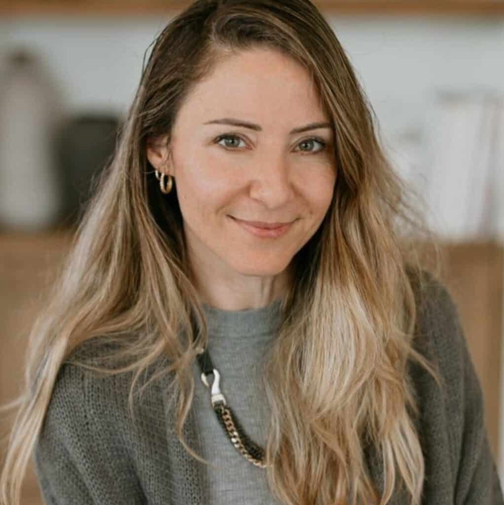 אישה עם שיער בלונדיני ארוך וגלי, לבושה בסוודר אפור ועונדת שרשרת, מחייכת למצלמה בתוך חלל סגור, מייצגת חום ותמיכה – בדיוק כמו הליווי להורים גרושים המבקשים הדרכה במסע ההורות שלהם.