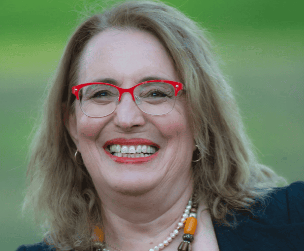 אישה עם שיער חום בהיר ומשקפיים אדומים מחייכת בחוץ, לבושה בחולצה כהה ועונדת שרשרת חרוזים צבעונית. דליה אוגמן מומחית ל–אימון גירושין וליווי גירושין, כאן כדי לתמוך בכם בתהליך הפרידה.