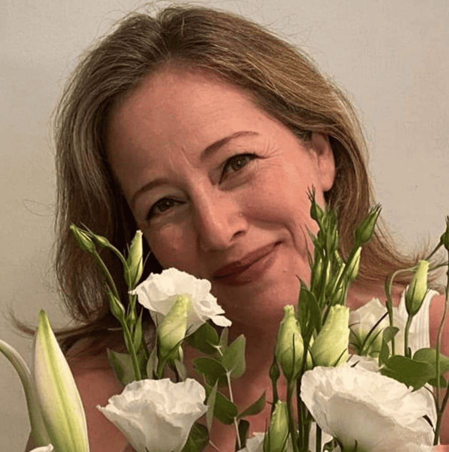 אישה עם שיער חום בהיר מחייכת מאחורי זר פרחים לבנים ועלים ירוקים, מעניקה רגע שמאפשר נשימה, על רקע אחיד. צילום: הדס זלצמן.