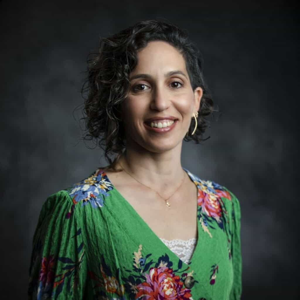 אישה עם שיער כהה ומתולתל, לבושה בשמלה פרחונית ירוקה ועגילי חישוק זהב, מחייכת למצלמה על רקע כהה ומטושטש, ומגלמת את הביטחון והחמימות הקשורים לליווי כלכלי של כרמי אור.