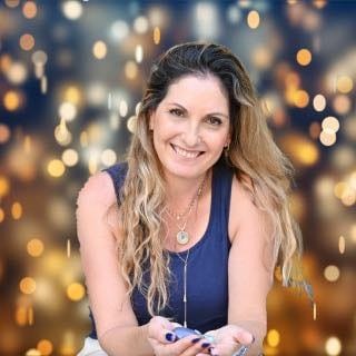 נומרולוגית ומאמנת NLP עם שיער ארוך וגלי, לבושה בחולצה ללא שרוולים, מחייכת למצלמה בזמן שהיא יושבת על רקע מטושטש עם אורות בוקה.