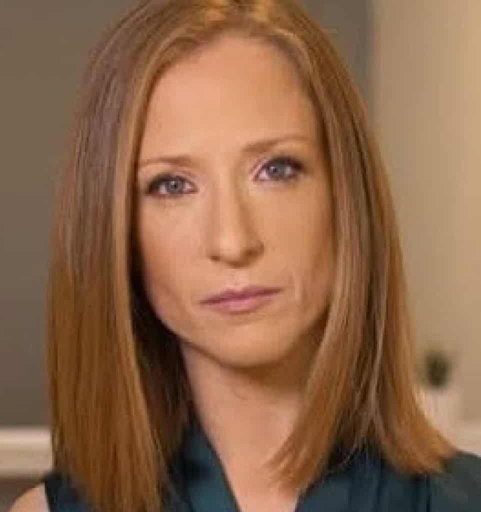 מנחת קבוצות הורים ומטפלת MSW עם שיער חום בהיר ישר באורך הכתפיים ועיניים כחולות מביטה ישירות למצלמה עם הבעה ניטרלית. הרקע מטושטש קלות.