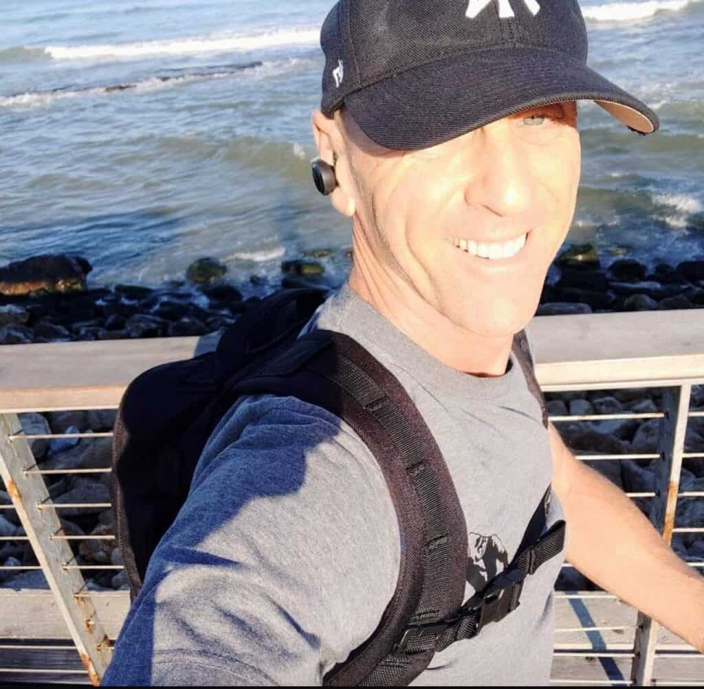 מאמן כושר איל בן זאב, לבוש בכובע שחור וחולצה אפורה, מצטלם סלפי ליד מעקה בטיילת עם גלים וסלעים ברקע – כי כושר ותודעה הולכים יחד גם בטבע.