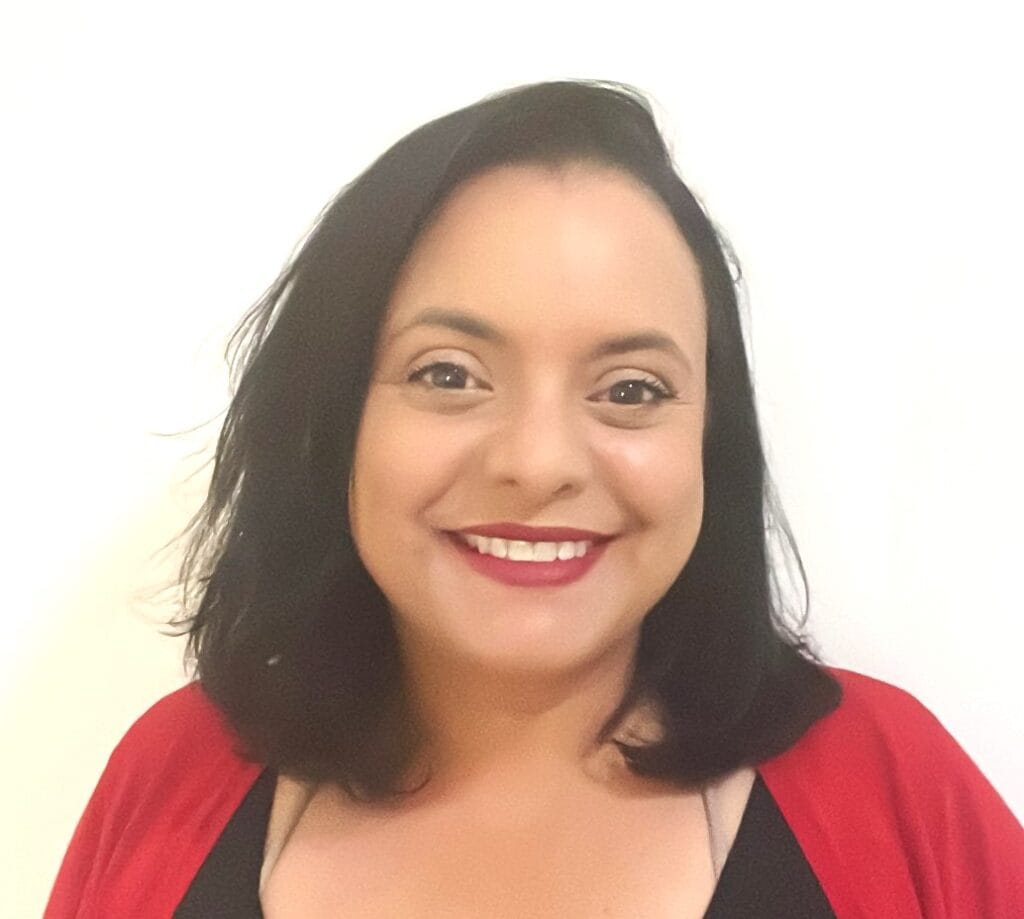 אישה עם שיער שחור באורך הכתפיים, לבושה בסוודר אדום וחולצה שחורה, מחייכת למצלמה על רקע לבן חלק. זו דיקלה אזולאי מדמון, מנחה למיניות בריאה ויועצת זוגית.