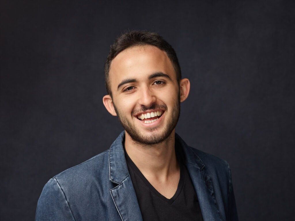 צעיר עם שיער קצר וכהה וזקן, לבוש ז'קט ג'ינס וחולצה שחורה, מחייך על רקע כהה וחלק — דניאל מור מומחה בטיפול רגשי וטיפול הוליסטי.