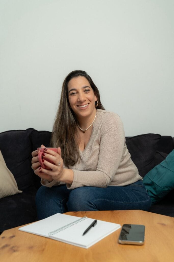 אישה יושבת על ספה ומחזיקה ספל, עם מחברת, עט וסמארטפון על השולחן לפניה — אולי מתכוננת לפגישה עם טל בלכר, יועצת זוגית.