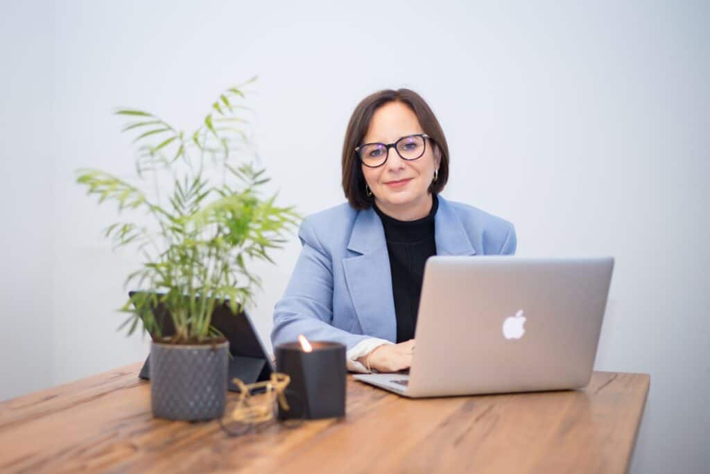 אקטוארית מיכל תמיר חייט, מרכיבה משקפיים ובלייזר כחול, יושבת ליד שולחן עץ עם מחשב נייד, צמח בעציץ, נר וטאבלט לפניה.
