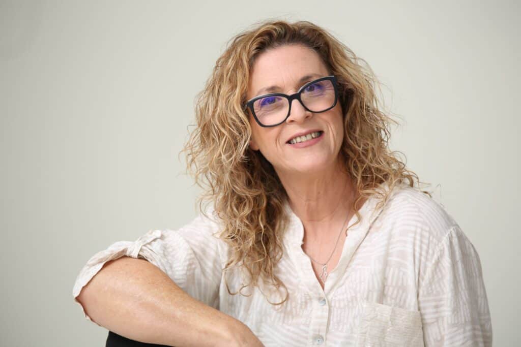 מאמנת נשים עם שיער בלונדיני מתולתל ומשקפיים כחולים, בחולצה בהירה, מחייכת קלות בישיבה מול רקע ניטרלי—מזמינה אותך לשיפור זוגיות ומערכות יחסים.