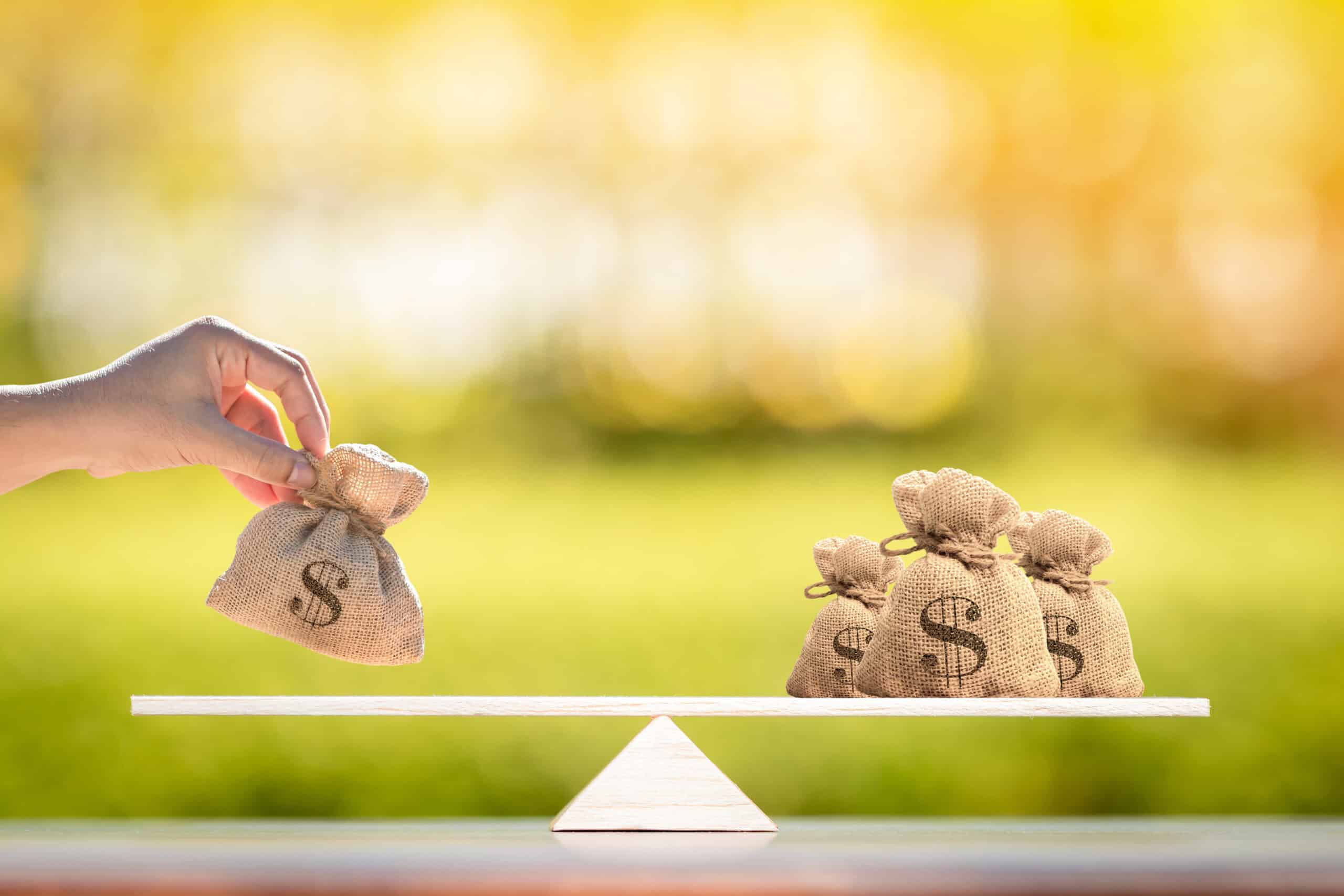 יד מניחה שקית כסף קטנה מבד יוטה על צד אחד של מאזניים מעץ, מול מספר שקיות דומות בצד השני, המסמלות איזון משאבים או פנסיוניים במהלך גירושים, על רקע חוץ מטושטש.