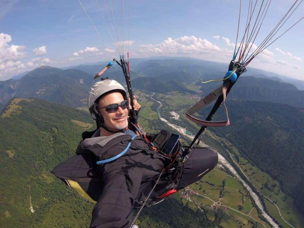 גבר גולש במצנח רחיפה גבוה מעל עמק ירוק עם גבעות, נהר מתפתל וכפרים פזורים למטה תחת שמים מעוננים חלקית, מחבק התחלה חדשה ואת תחושת החופש שבני בן ישראל מוקיר.