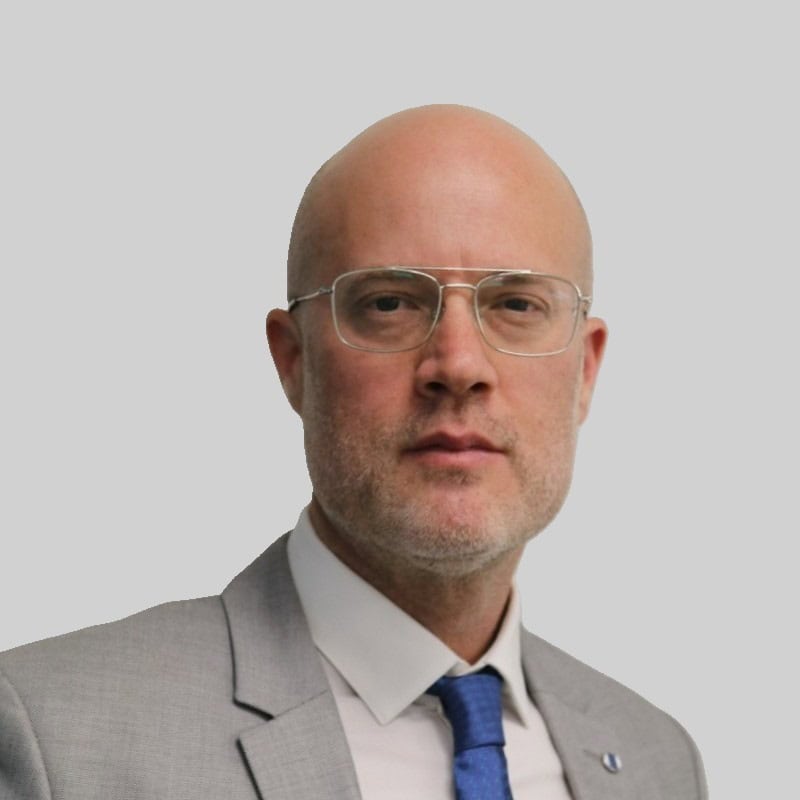 גבר קירח עם משקפיים שקופים, לבוש בחליפה אפורה ועניבה כחולה, מביט ישירות למצלמה על רקע בהיר וחלק.