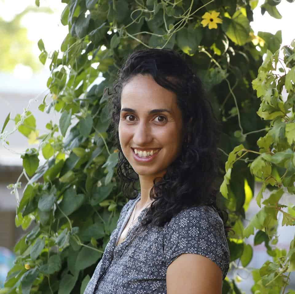 מירב ארווס, אישה עם שיער כהה מתולתל ולבושה בחולצה עם דוגמה, ניצבת מול צמחיה ירוקה ומחייכת למצלמה, במהלך מפגשי תנועה באווירה טבעית.