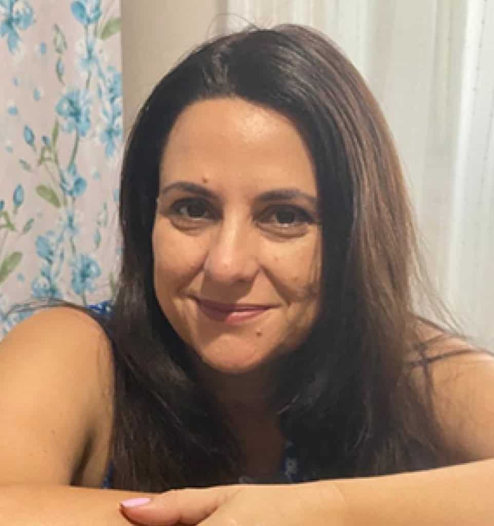 אישה עם שיער ארוך וכהה יושבת בתוך הבית ומחייכת למצלמה. וילונות לבנים ובד עם דוגמת פרחים ברקע יוצרים אווירה חמימה המתאימה למדריכת הורים התומכת בהורים גרושים.