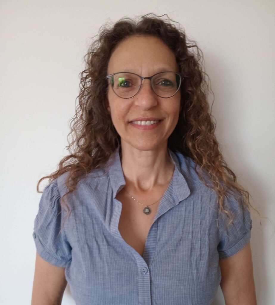 יועצת לכלכלת משפחה מחייכת, עם שיער חום מתולתל, משקפיים וחולצה כחולה קצרה, עומדת על רקע לבן חלק, מקרינה חום וביטחון.