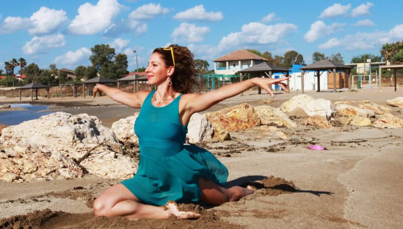 אישה בשמלה בצבע טורקיז יושבת על החול בתנוחת פיסוק עם זרועות מושטות, ליד סלעים גדולים על חוף תחת שמיים מעוננים חלקית, ומפגינה גמישות בהשראת דידה פיטנס.