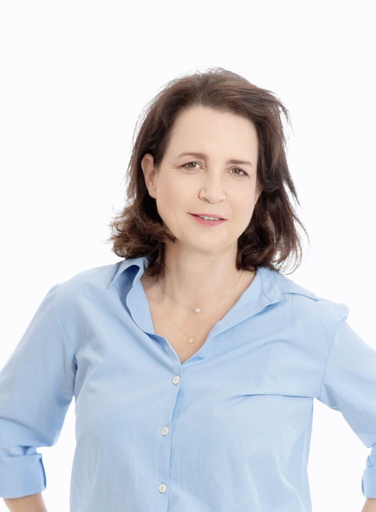 קארין זרעוני, מאמנת אישית, עם שיער חום באורך הכתפיים לובשת חולצה כחולה בהירה מכופתרת, ניצבת מול רקע לבן נקי.
