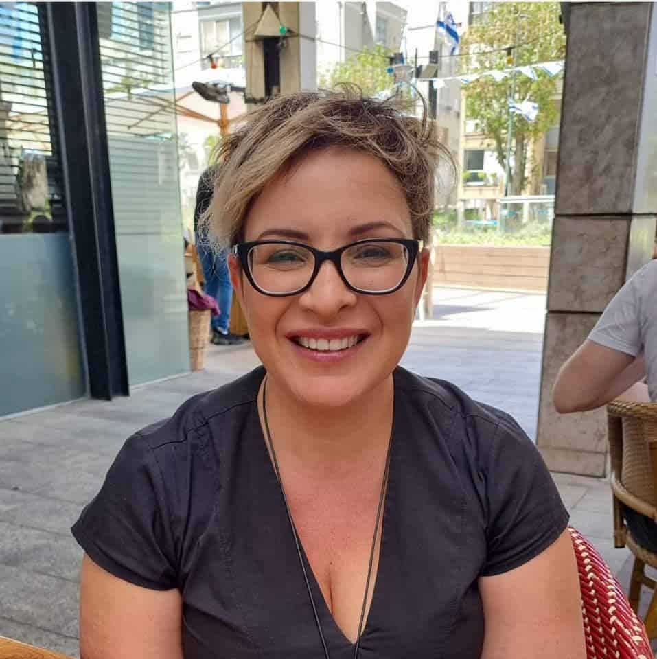 אישה עם שיער בלונדיני קצר ומשקפיים יושבת בשולחן בית קפה בחוץ, מחייכת למצלמה. ברקע נראים בניינים עירוניים וצמחייה. קרן אור קמלמן, מטפלת CBT ופסיכולוגית חינוכית, מציגה אווירה נעימה ובטוחה.