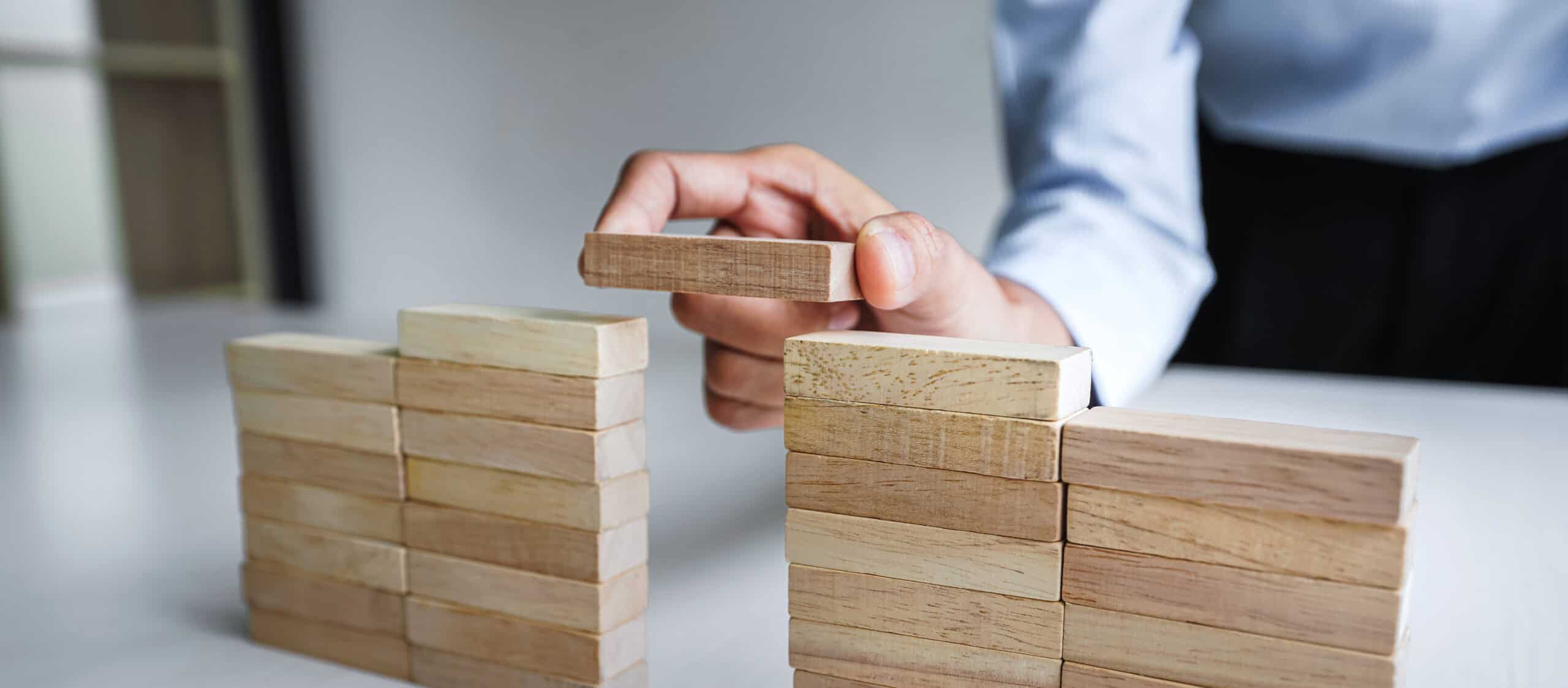 יד של אדם מניחה קוביית עץ כדי לגשר על פער בין שתי ערימות קוביות עץ על שולחן לבן, המסמל את השאלה: האם מגשר/ת לגרושין חייב/ת להיות עורך דין?