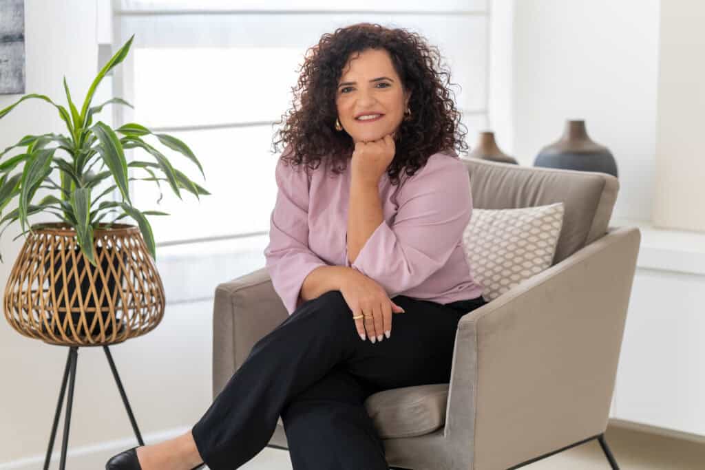 אישה עם שיער מתולתל יושבת בכורסה ומחייכת למצלמה. לבושה בחולצה סגולה בהירה ומכנסיים שחורים, היא מקרינה חום – ומבטאת את מהות מעגלים של תמיכה – עם צמח ואגרטלים דקורטיביים ברקע.