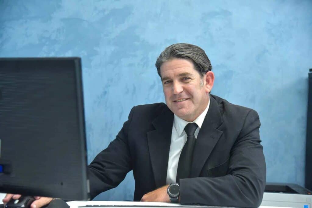 גבר בחליפה כהה ועניבה יושב ליד שולחן, מחייך למצלמה, מולו מסך מחשב וברקע קיר כחול, המרמז על מומחיותו בדיני משפחה או ירושה.