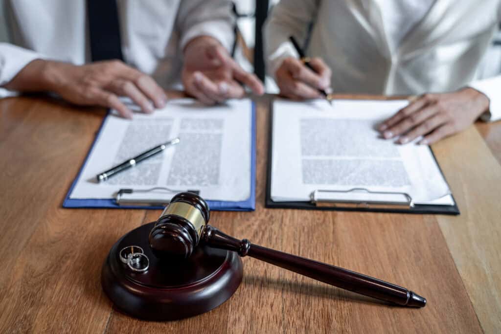 שני אנשים לבושים בבגדי עסקים יושבים ליד שולחן וחותמים על מסמכים, עם פטיש ושתי טבעות נישואין על גוש עץ בקדמת התמונה — תמונה המשקפת את גירושין — איך נפרדים יפה?