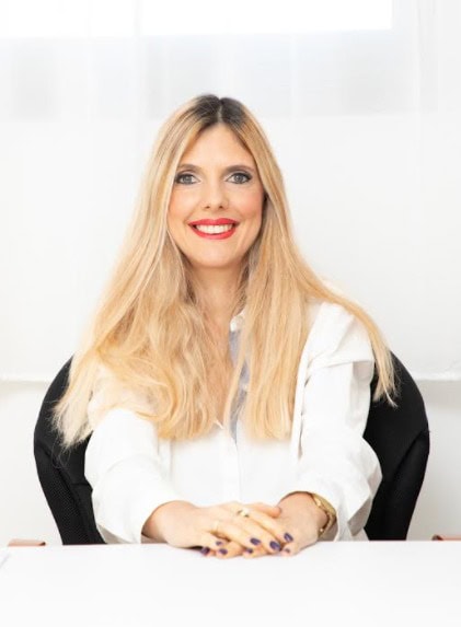 אישה עם שיער בלונדיני ארוך, לבושה בחולצה לבנה, יושבת ליד שולחן ומחייכת למצלמה — עורכת דין המתמחה בדיני משפחה.