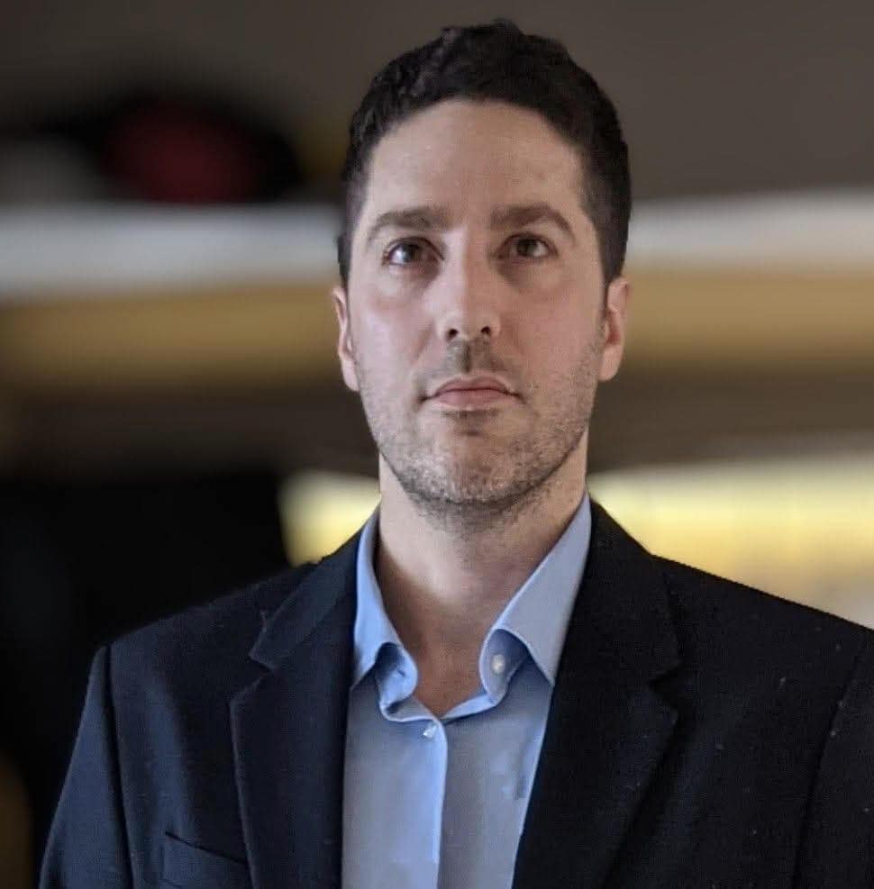 גבר עם שיער קצר וכהה, לבוש בז'קט כהה וחולצה תכלת, עומד בתוך מבנה על רקע מטושטש במועדון החברים הפיננסי, המתמחה בתכנון פיננסי.