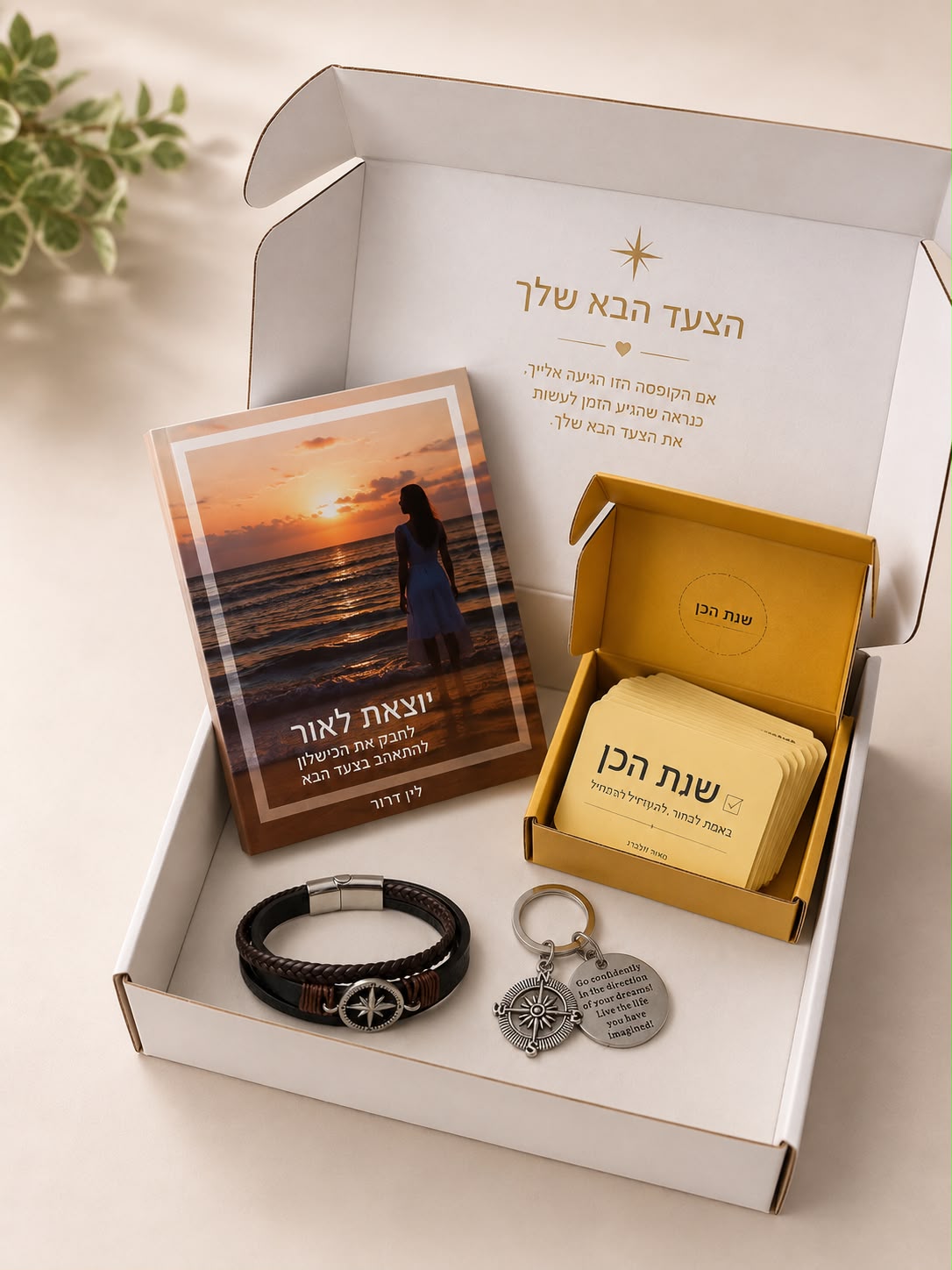 קופסת מתנה פתוחה המכילה ספר בעברית בשם “הצעד הבא שלך”, קופסת כרטיסים צהובה, צמיד קלוע ושני מחזיקי מפתחות ממתכת עם תליונים וכיתוב חרוט, כולם מסודרים בקפידה על משטח ניטרלי.