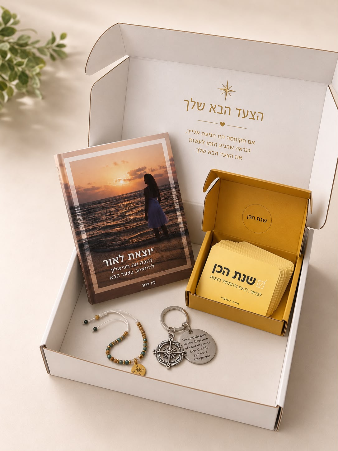 קופסת מתנה ובה ספר בעברית, סט כרטיסים בקופסה צהובה שעליה הכיתוב “כאן בפנים”, מחזיק מפתחות מכסף וצמיד חרוזים — כולם מסודרים בקפידה על משטח בהיר כדי לעורר השראה לנקוט בצעד הבא.